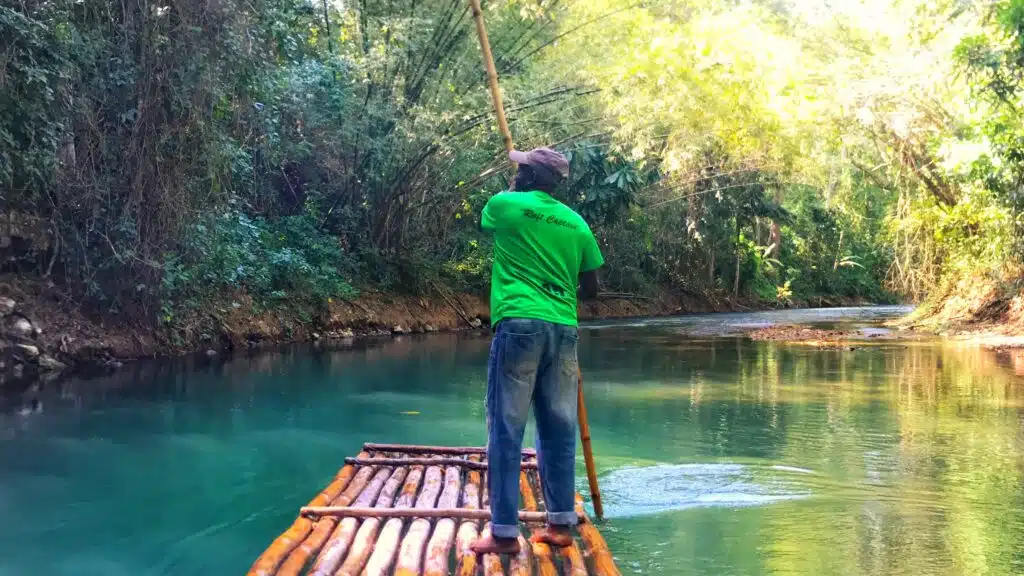 A raft captain in a bright green shirt guides a bamboo raft down the calm, emerald waters of the Martha Brae River in Trelawny, Jamaica, surrounded by lush tropical greenery and golden sunlight filtering through bamboo