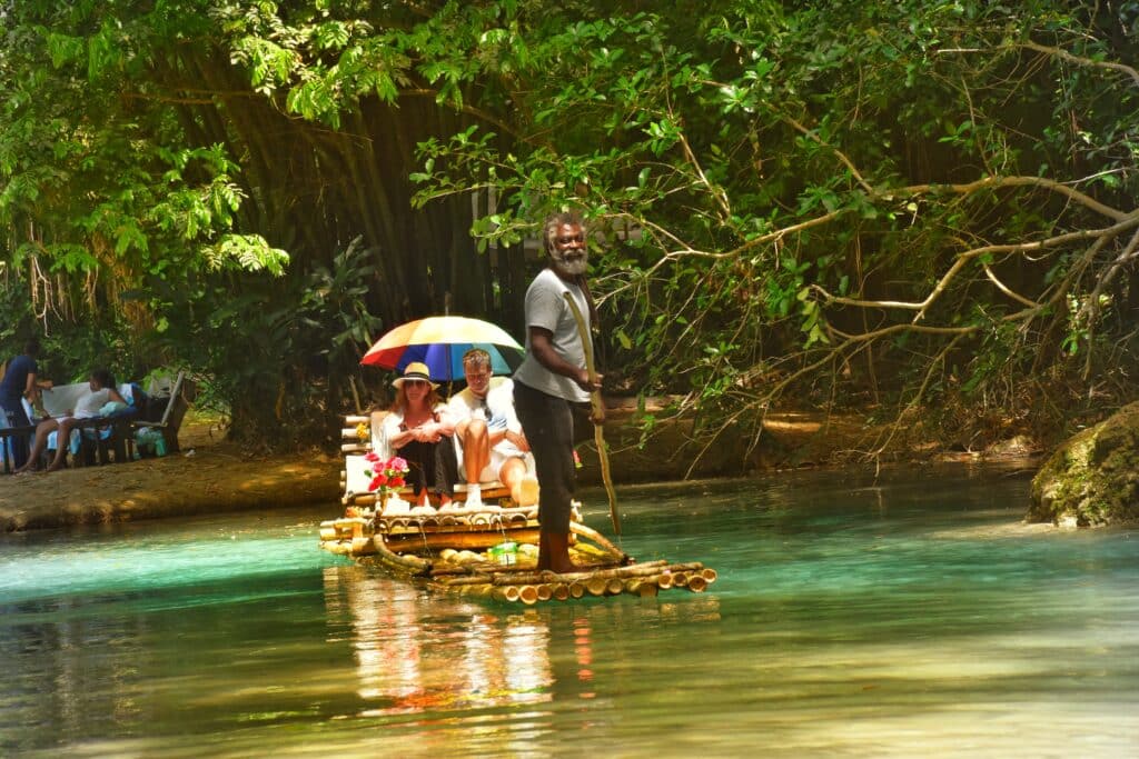 A Jamaican raft captain guides two visitors down the clear turquoise waters of the White River in Ocho Rios, Jamaica, surrounded by lush tropical greenery — a popular spot for bamboo rafting, swimming, and relaxation