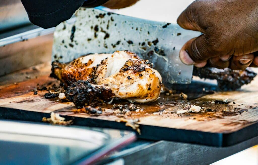 Jamaican jerk chicken being chopped on a wooden board at a roadside jerk pan
